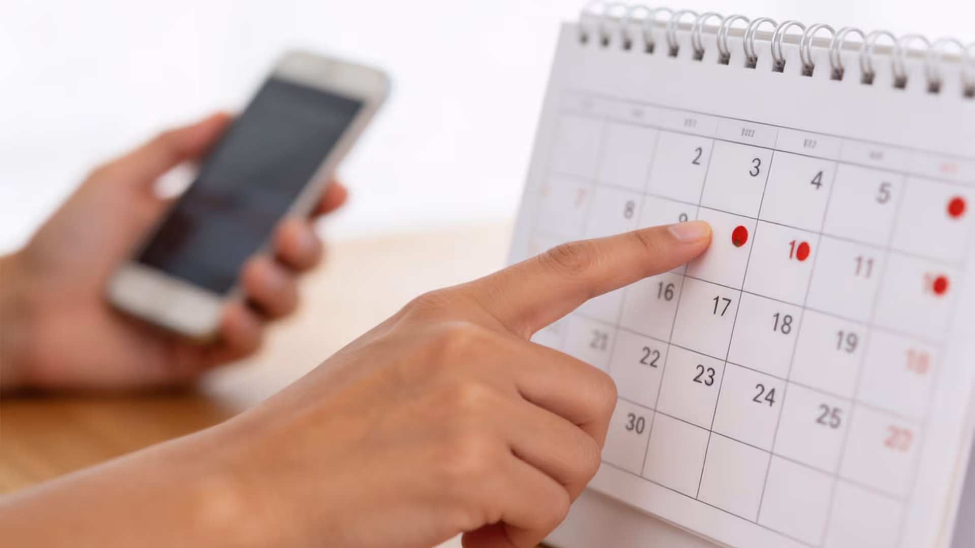 Mano señalando una fecha marcada con punto rojo en un calendario de escritorio junto a un celular.