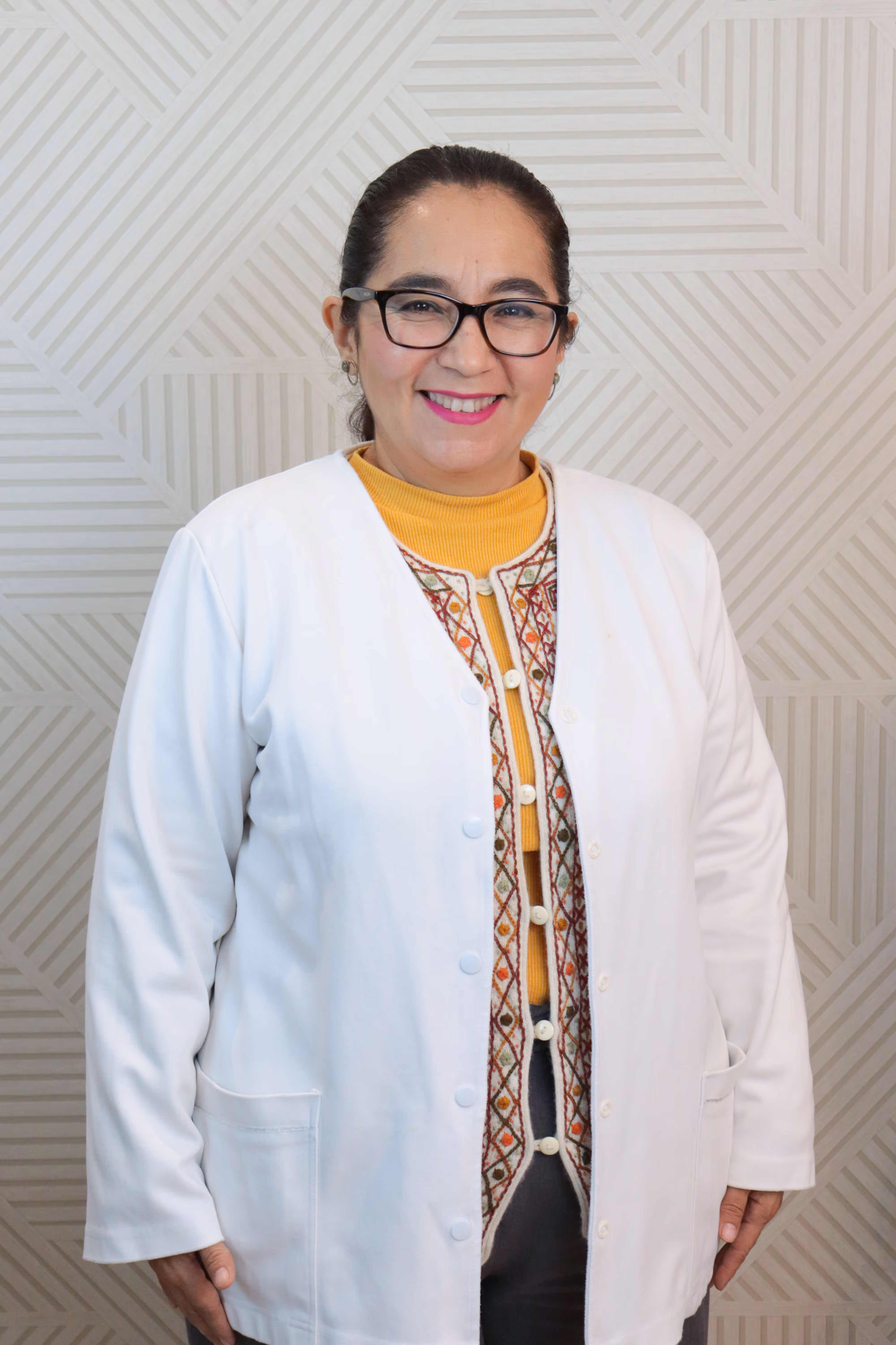 Mujer sonriente con gafas y bata blanca sobre un suéter amarillo y chaleco bordado.