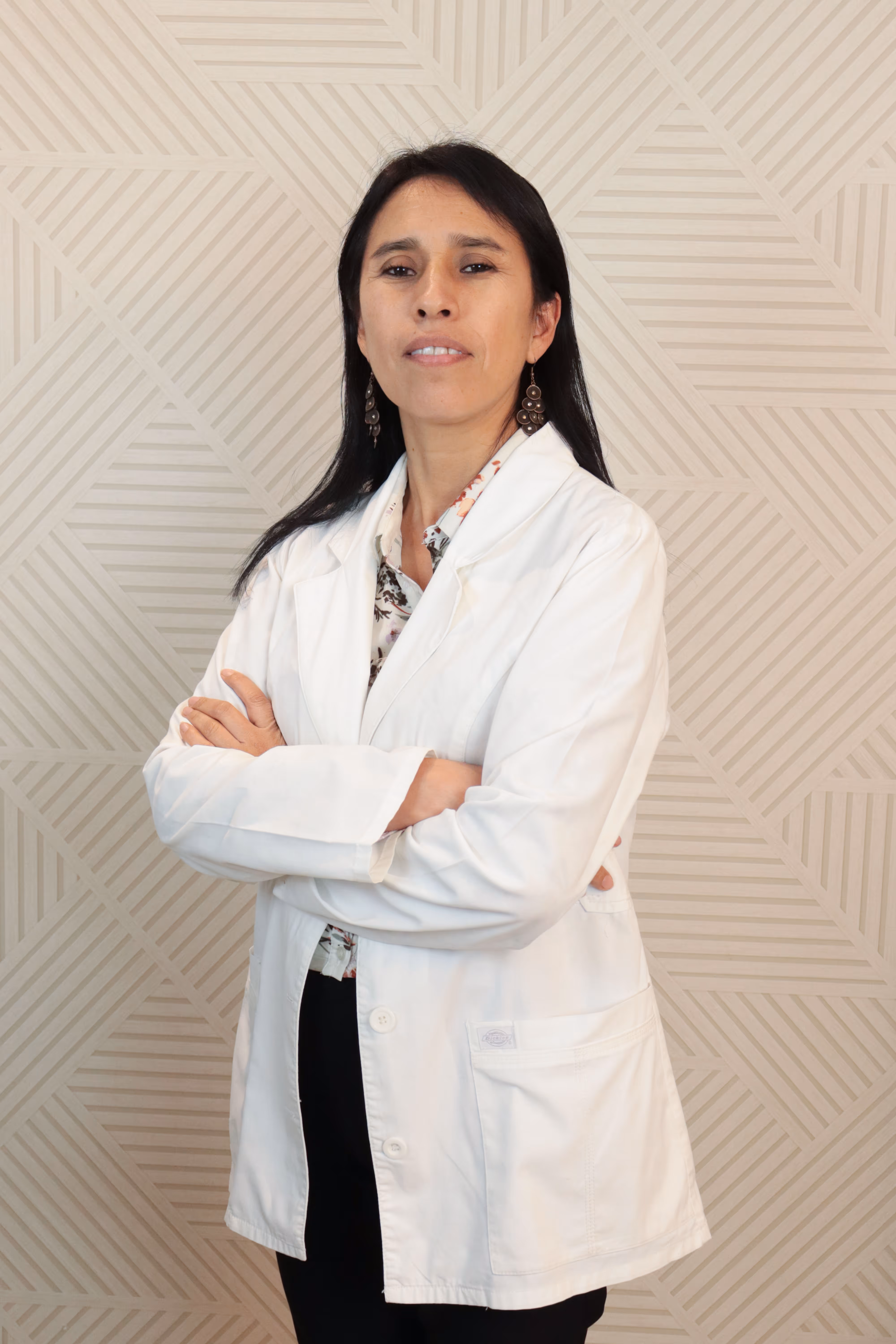 Mujer con cabello negro y largo, vestida con bata blanca, posa con los brazos cruzados frente a una pared con patrón geométrico.