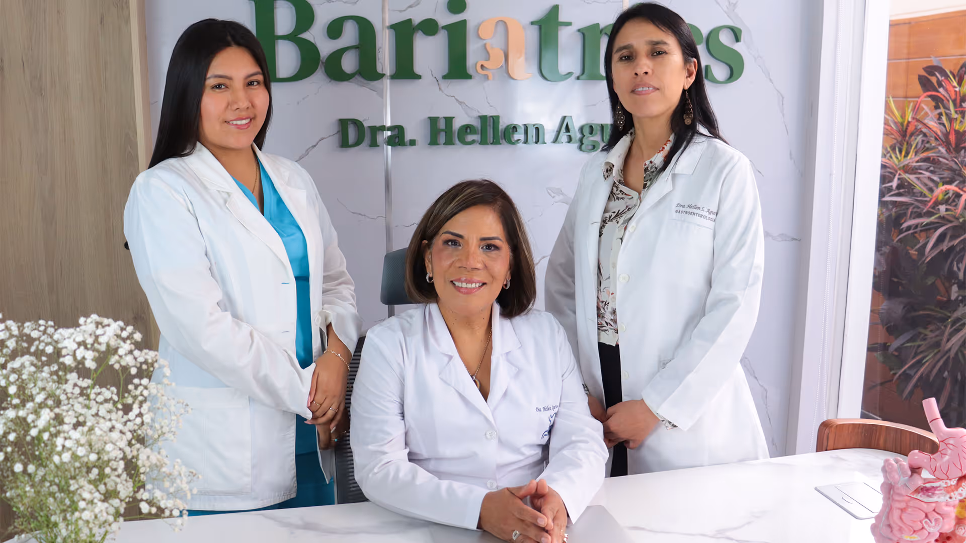 Tres médicas sonrientes con batas blancas en una consulta de bariatría, con un modelo anatómico del sistema digestivo en la mesa.