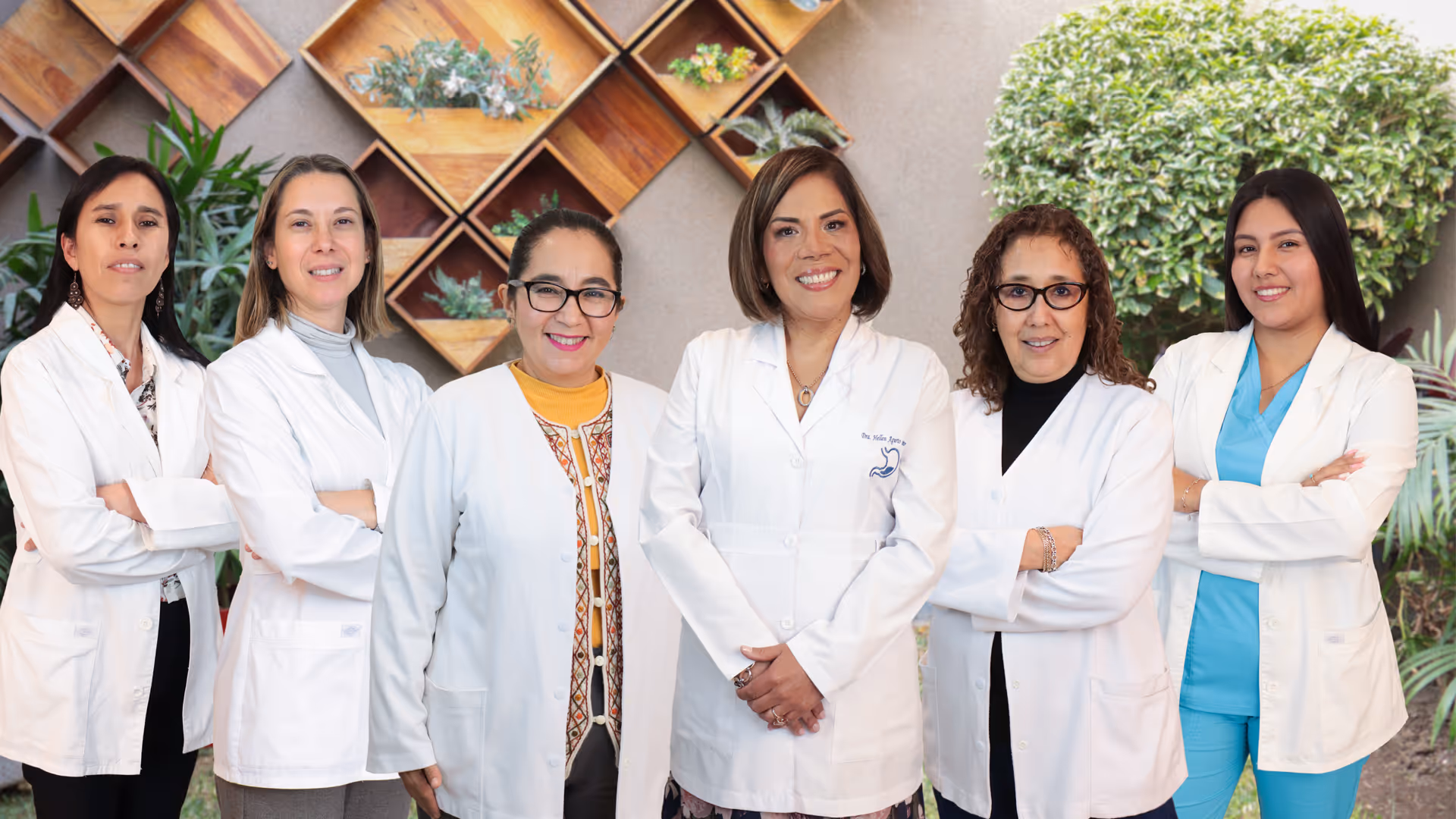 Seis mujeres profesionales con batas blancas posan juntas frente a una pared decorada con plantas.